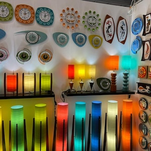 May include: A wall display of colorful mid-century modern decor, including clocks, lamps, and wall art. The clocks feature various designs and colors, including blue, green, and orange. The lamps are made of wood and have colorful shades. The wall art features abstract designs and geometric shapes.