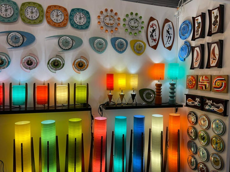 May include: A wall display of colorful mid-century modern decor, including clocks, lamps, and wall art. The clocks feature various designs and colors, including blue, green, and orange. The lamps are made of wood and have colorful shades. The wall art features abstract designs and geometric shapes.