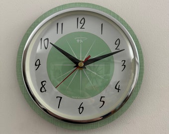 May include: A circular wall clock with a silver-coloured bezel and a light green face. The clock features black hands, black numerals, and a red second hand. The clock face has a light green compass design in the centre. The clock is labelled "MADE IN ENGLAND".
