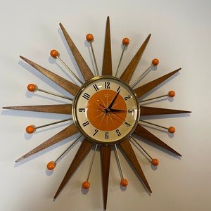 Puede incluir: Un reloj de pared de estilo moderno de mediados de siglo con un diseño de sol. El reloj tiene un marco de madera con acentos naranjas y una cara blanca con números negros.