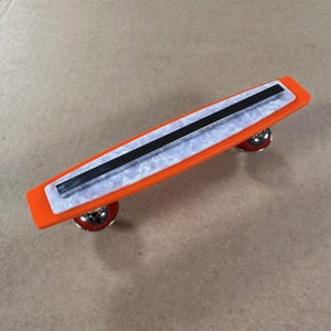 May include: An orange skateboard-shaped brooch with a white and pearlescent center and a black stripe. The brooch has two silver wheels with red accents. A unique accessory.