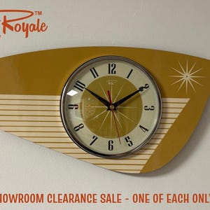 May include: A mustard yellow wall clock with an asymmetrical, elongated shape. The clock face features a cream-coloured centre with black numerals, black hour and minute hands, a red second hand, and a white starburst graphic on the right side.