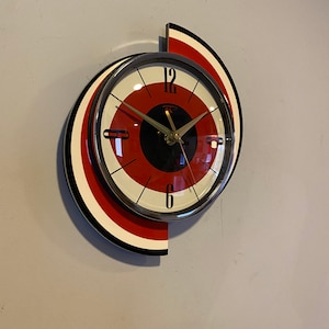 May include: A retro-style wall clock with a red, black, and white design. The clock face features a red and white target pattern with black accents and gold hands. The clock is mounted on a wall, showcasing its unique, mid-century modern aesthetic.
