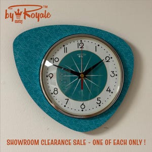 May include: A teal wall clock with an asymmetrical, rounded triangular frame featuring a white, swirling line pattern. The clock face has a white background with black numerals, a dark teal centre, black hour and minute hands, and a red second hand.