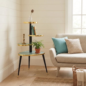 May include: A three-tiered plant stand with a black central pole and gold-toned trim. The shelves are a light blue-green color. The stand has three black legs with gold-toned feet. A small potted plant, books, and a candle holder are on the shelves.