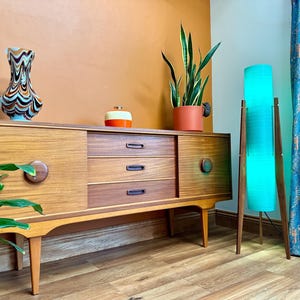 Könnte beinhalten: Ein modernes Sideboard aus der Mitte des Jahrhunderts mit drei Schubladen und zwei Türen. Das Sideboard ist aus Holz und hat eine hellbraune Oberfläche. Es steht auf einem Holzboden. Im Hintergrund befinden sich eine Vase mit einem wirbelnden Muster, eine Topfpflanze und eine Stehlampe mit einem türkisfarbenen Schirm.