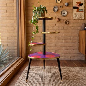 Puede incluir: Un soporte para plantas de varios niveles con un poste central negro y cuatro estantes circulares. Los estantes tienen bordes dorados y una superficie morada con un patrón floral rojo y naranja. El soporte se apoya sobre tres patas negras.
