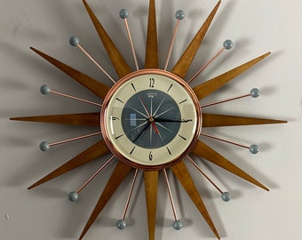 May include: A mid-century modern wall clock with a sunburst design. The clock has a wooden frame with copper-colored accents and a gray face with black hands. The clock has a diameter of approximately 12 inches.