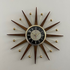 May include: A mid-century modern wall clock with a black face and gold accents. The clock has a starburst design with wooden rays and gold-toned metal balls at the ends of each ray.