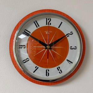 May include: A round wall clock with a bright orange outer rim featuring a textured pattern and a chrome border. The clock face has a white background with black numerals, a central orange circle with white starburst lines, and black hands.