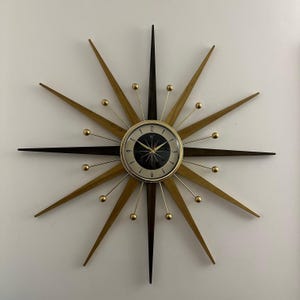 Könnte beinhalten: Eine Mid-Century-Modern-Sternenburst-Uhr mit goldenen und braunen Holzstrahlen und einem schwarzen Zifferblatt. Die Uhr hat goldene Zeiger und goldene Akzente um das Zifferblatt.