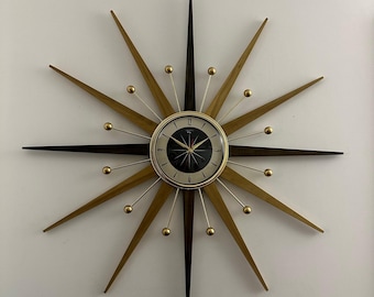 May include: A mid-century modern starburst clock with gold and brown wooden rays and a black clock face. The clock has gold hands and gold accents around the clock face.