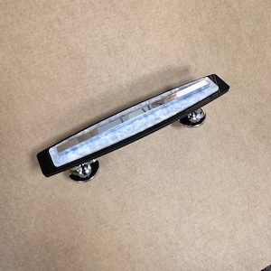 May include: A black cabinet pull with a rectangular design. The centre features a white and blue marbled pattern with a silver accent. The pull is mounted on two round, silver-coloured knobs.
