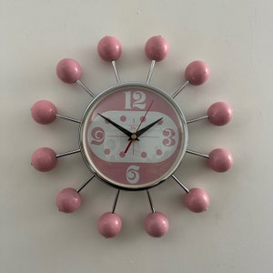 May include: A pink and silver clock with a white face and black hands. The clock has a unique design with pink balls around the edge.
