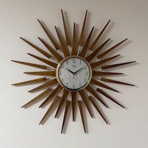 May include: A sunburst wall clock featuring a circular white face with black Arabic numerals and a gold-toned metal rim. The clock is surrounded by numerous pointed wooden rays extending outwards in a starburst pattern with black clock hands.