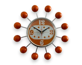 May include: A mid-century modern wall clock with an orange face and silver accents. The clock has a unique design with 12 orange spheres around the clock face.