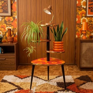 May include: A vintage plant stand with a black central pole, gold trim, and a patterned orange and red surface. It has three circular shelves holding potted plants and a small desk lamp. The stand's base is triangular, with black legs.