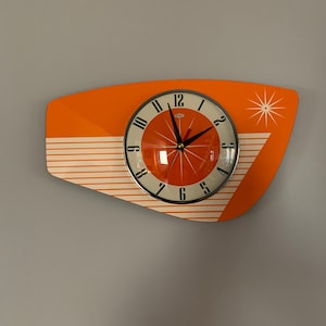 Puede incluir: Reloj de pared moderno de mediados de siglo, naranja y blanco, con una esfera negra y agujas plateadas. El reloj tiene un diseño de estrella y un patrón de rayas blancas.