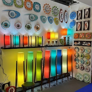 May include: A wall display of colorful mid-century modern lamps and clocks. The lamps are made of woven bamboo and come in a variety of colors, including yellow, green, blue, and red. The clocks are all round and have a variety of designs, including abstract patterns and geometric shapes.