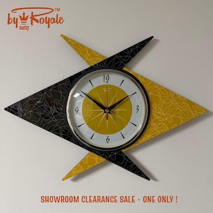 May include: A starburst wall clock featuring a central circular face with a yellow centre, white outer ring, and black numerals. The clock is mounted on a diamond-shaped backing with a black and yellow pattern of intersecting white lines.