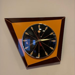 May include: A modern wall clock with a black face and orange and brown trim. The clock has a unique design with a triangular shape and a silver frame.
