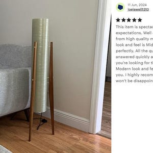 May include: A tall, cylindrical floor lamp with a white, textured shade and a wooden tripod base. The lamp is standing on a wooden floor next to a gray armchair.