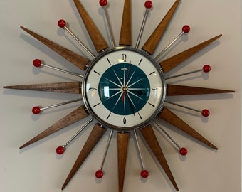 Puede incluir: Un reloj de pared de estilo moderno de mediados de siglo con un diseño de estrella. El reloj tiene una esfera azul verdosa con números blancos y detalles rojos. El reloj está hecho de madera y metal.