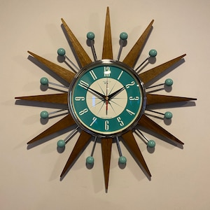 May include: A teal and white clock with a starburst design. The clock has a silver frame and black hands. The starburst design is made of wood and has teal accents.