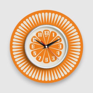 Peut inclure: Horloge murale orange et blanche avec un motif en forme de soleil. Le cadran de l'horloge a un fond blanc avec des chiffres orange et un cadre argenté.