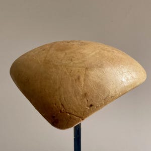 May include: A light brown, rounded wooden hat block with a smooth, polished surface and visible natural wood grain patterns. The object is mounted on a thin, dark vertical metal stand, showcasing a curved, dome-like shape used for millinery.