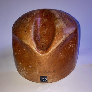 Op de afbeelding: Een bruine houten hoedenblok met een gladde, ronde bovenkant en een duidelijke centrale inkeping. Het oppervlak vertoont natuurlijke houtnerven, kleine krasjes en gebruikssporen. Een zwart label met het witte cijfer 55 is bevestigd.