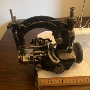 Op de afbeelding: Een vintage naaimachinemechanisme van zwart metaal met zichtbare tandwielen, een gebogen arm en een garenkloshouder. Een houten liniaal ligt eronder met metingen in centimeters, waarbij de machine zich uitstrekt van 86 tot 117 cm.