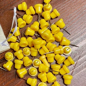 May include: A pile of yellow plastic push pins with metal points. The push pins are scattered on a brown wooden surface.