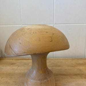 May include: A wooden hat block shaped like a mushroom. The light-coloured wood hat block has a rounded top and a pedestal base. The wood grain is visible. It is on a wooden surface.