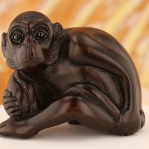 May include: A dark brown wooden carving of a monkey sitting with its arms crossed and holding a round object.