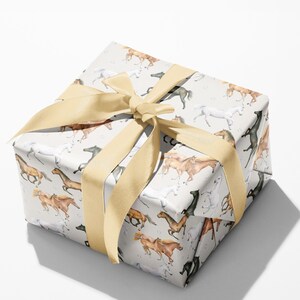 May include: A wrapped gift box featuring a light gray paper with a repeating pattern of running horses in various colors. The box is tied with a wide, light gold satin ribbon, forming a large bow on top. The gift wrap is suitable for any occasion.