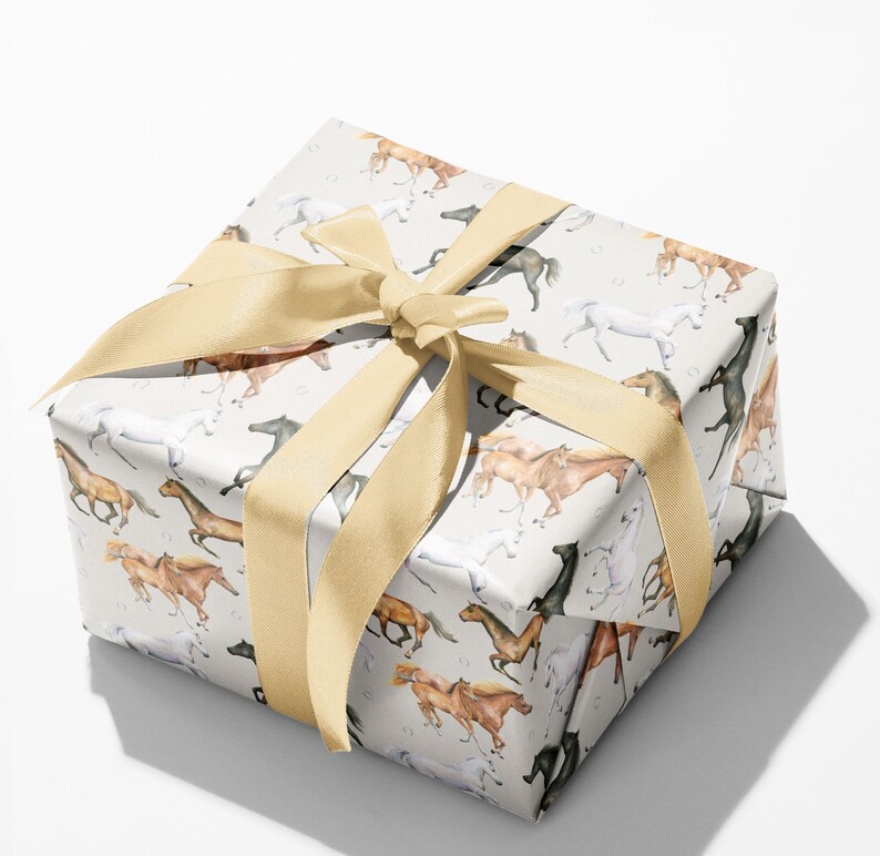 May include: A wrapped gift box featuring a light gray paper with a repeating pattern of running horses in various colors. The box is tied with a wide, light gold satin ribbon, forming a large bow on top. The gift wrap is suitable for any occasion.