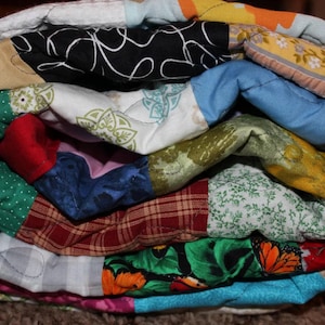 May include: A colorful patchwork quilt with various patterns and fabrics, including polka dots, floral prints, and plaid. The quilt is folded and stacked, showcasing the different layers and textures.