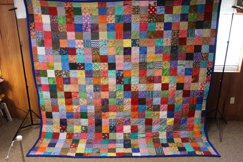 May include: A large quilt made of many colorful fabric squares, each with a different pattern. The quilt is bordered with a dark blue fabric.