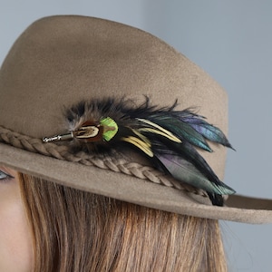Puede incluir: Sombrero fedora marrón con una banda trenzada y plumas decorativas. Las plumas son una mezcla de verde, negro y azul iridiscente. La persona que lleva el sombrero tiene el pelo largo y castaño claro.