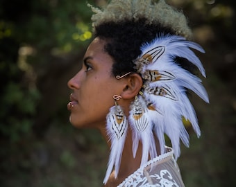 May include: A woman with short, blonde hair wearing a white lace top and a white feather headdress with brown accents. She is wearing a pair of feather earrings with brown accents.
