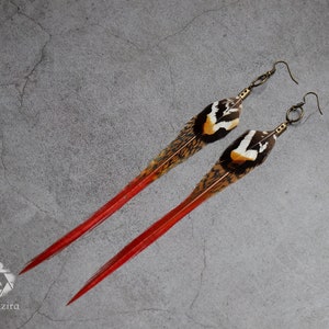 Puede incluir: Un par de pendientes largos de plumas con puntas rojas. Las plumas son marrones, blancas y amarillas con un diseño estampado. Los pendientes están hechos con ganchos de bronce.