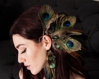 Puede incluir: Un pendiente de plumas de pavo real con un marco de alambre dorado. Las plumas están dispuestas en cascada con colores verde, azul y marrón.