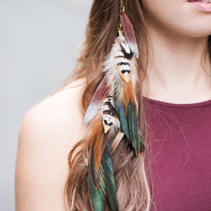 Könnte beinhalten: Eine Frau mit langen braunen Haaren trägt ein burgunderfarbenes Oberteil mit einer Federhaarextension. Die Extension besteht aus braunen, grünen und weißen Federn und ist an einer goldenen Kette befestigt.