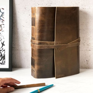 May include: A brown leather-bound journal with a tan leather strap and buckle closure. The journal has a distressed look and is embossed with the word "EGCHOL".