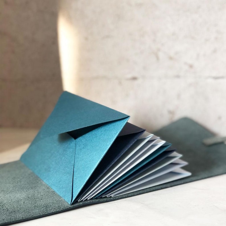 May include: A teal-colored, handmade paper booklet with a folded cover, fanned open to reveal multiple pages. The booklet rests on a gray leather-like surface, suggesting a journal or notebook.