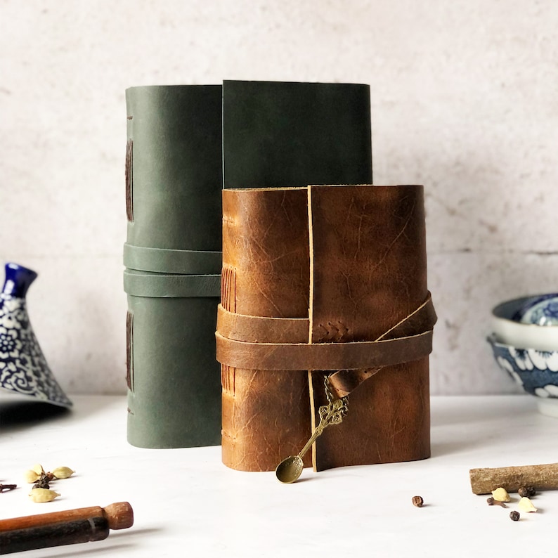 May include: Two leather-bound journals, one green and one brown, with a decorative metal spoon charm attached to the brown journal.
