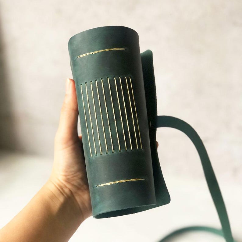 May include: A dark teal leather journal with gold stitching. The cylindrical journal is held by a green metal stand. The journal has gold stitching along the spine and at the top and bottom.
