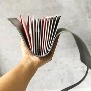 May include: A gray leather wallet with multiple card slots, fanned open to display colorful cards. The wallet is held in a hand against a neutral background. The cards have red, pink, and white edges.