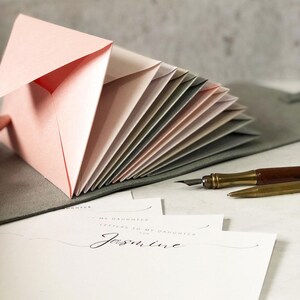 May include: A collection of pink and neutral-toned envelopes fanned out, with a handwritten letter that reads "Letters to My Daughter for Jasmine." A vintage pen and pencil are also visible.